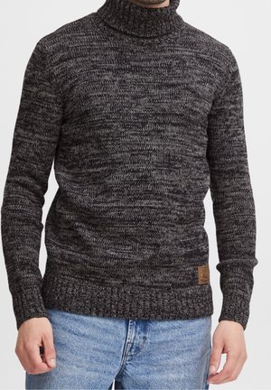 Homme portant un pull en tricot col roulé noir et gris chiné avec une pièce en cuir en bas et un jean bleu clair.