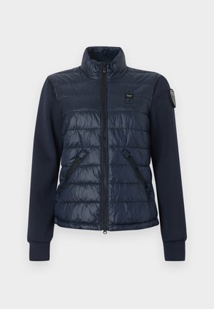 Γυναικείο υβριδικό μπουφάν σε ναυτικό μπλε με καπιτονέ μπροστινό μέρος, μανίκια από ομαλό ύφασμα, τσέπες με φερμουάρ, ψηλό γιακά και επώνυμα μπαλώματα στο στήθος και το μανίκι.