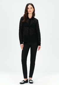 Camicia nera in pizzo con bottoni e maniche lunghe, dotata di colletto e una texture trasparente. Abbinata a pantaloni neri e scarpe bicolore.
