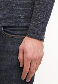 Mörk heathergrå l ångärmad skjorta med texturerad tyg och en liten logotagg nära nederkanten, tillsammans med mörkblå jeans.