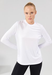 Langermet hvit treningstopp i fukttransporterende stoff, rund hals, sidesplitt, glatt tekstur, tettsittende design, kombinert med svarte leggings.