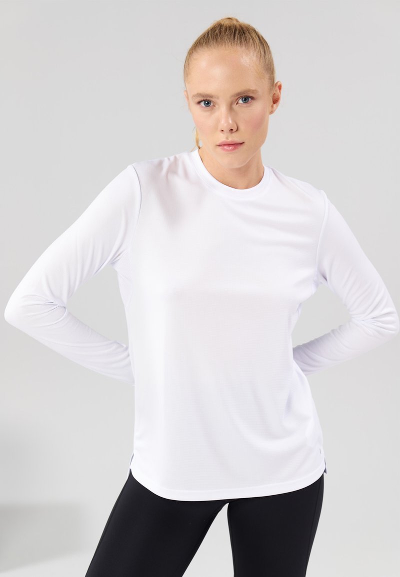 Langermet hvit treningstopp i fukttransporterende stoff, rund hals, sidesplitt, glatt tekstur, tettsittende design, kombinert med svarte leggings.
