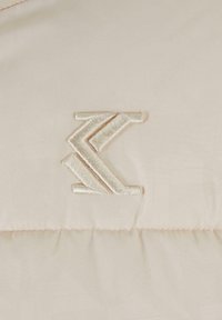 Beige pufferjakke i stoff med et relieff, lysfarget logo-design. Myk tekstur og subtilt sydd, med en matt finish.
