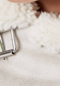 Kremowa wełniana tkanina o puszystej fakturze, wyposażona w srebrzystą klamrę i miękkie, zaokrąglone dodatki w widoku z bliska.