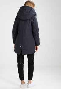 Długa ciemna kurtka z kapturem, ściągaczem w talii i zatrzaskami z tyłu. Ma gładką fakturę i jest zestawiona z czarnymi spodniami oraz białymi butami.