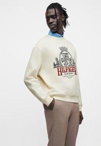 Εκρού φούτερ με μαύρο και κόκκινο τυπωμένο λογότυπο, με μοτίβο λιονταριού με κορόνα· συνδυασμένο με ανοιχτό καφέ παντελόνι, με απαλή υφή υφάσματος.