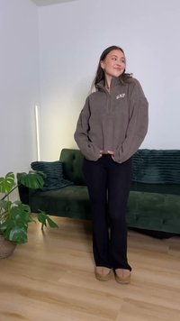 Maglione marrone in pile con mezza cerniera, con le lettere "EAP" sul petto. Pantaloni neri svasati e scarpe beige senza lacci. Divano verde sullo sfondo.