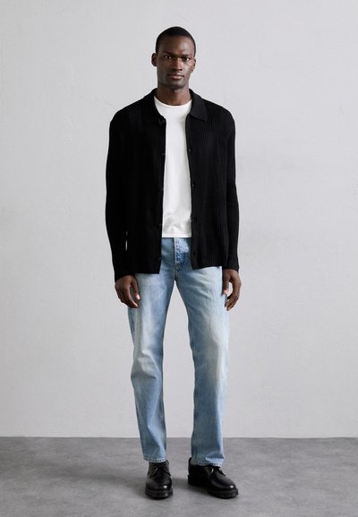 Svart ribbad cardigan, vit t-shirt, ljusblå jeans och svarta skor. Modellen står mot en neutral bakgrund med ett neutralt uttryck.