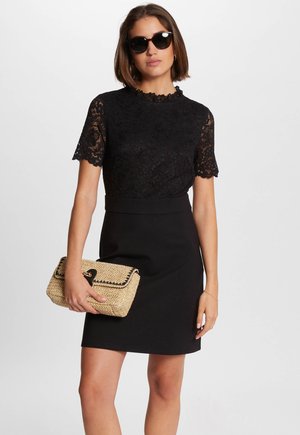 Black lace top with scalloped edges, paired with a fitted black skirt. Holding a beige woven clutch with black trim and a metal clasp.
