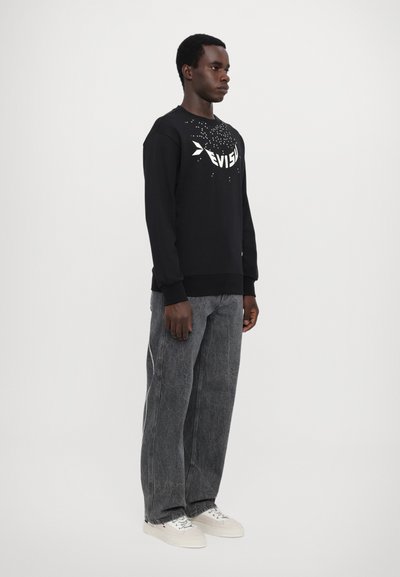 Sweatshirt noir avec logo blanc et design éclaboussé, associé à un jean gris baggy et des baskets blanches. Modèle debout contre un fond uni.