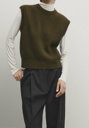Personne portant un pull sans manches en maille côtelée vert olive sur un haut blanc à manches longues avec un pantalon noir taille haute.