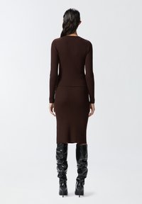 Vestito a maglia a coste marrone con maniche lunghe, silhouette aderente, orlo fino al ginocchio. Indossato con stivali neri al ginocchio, texture liscia, design minimale.