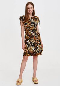 Schwarzes Kleid mit einem tropischen Blättermuster in Grün, Orange und Weiß, seitlichen Bindebändern und taillierter Taille, kombiniert mit geflochtenen Plateauschuhen.