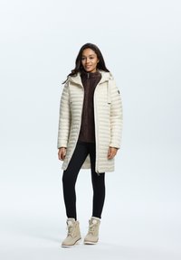 Lange crème pufferjas met een capuchon, ritssluiting aan de voorkant en een gebreid ontwerp onder een trui. Zwarte leggings en beige sneeuwlaarzen met vachtaccenten.