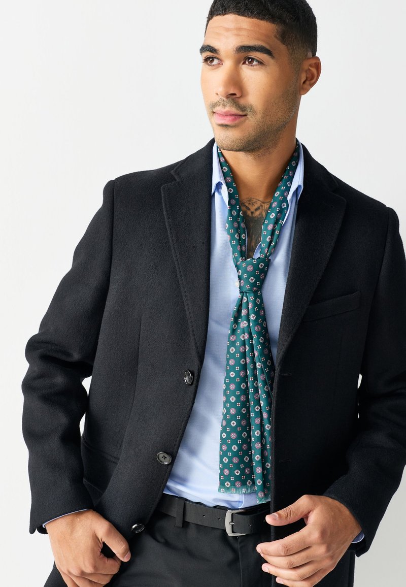 Black wool blazer with a tailored fit, worn over a light blue shirt. A green patterned silk scarf is draped around the neck.