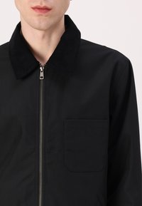 Chaqueta negra con cremallera, hecha de una tela suave y con un cuello de pana, que cuenta con un bolsillo en el pecho y un diseño minimalista.