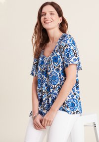 Bluse mit Blumenmuster, die blaue und schwarze Designs auf einem weißen Hintergrund zeigt, mit einem großzügigen Ausschnitt und kurzen Ärmeln. Weicher Stoff.