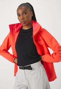 Veste imperméable rouge vif avec un col haut, des poignets élastiques, une doublure intérieure contrastante, portée sur une chemise noire et un pantalon de couleur claire.