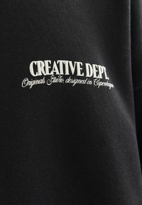 Zwarte sweatshirt met witte cursieve borduursel met de tekst "CREATIVE DEPT. Originals Studio, ontworpen in Kopenhagen." Zacht, gestructureerd materiaal.