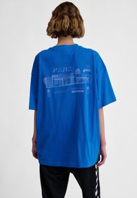 Överdimensionerad blå t-shirt i bomull med en grafisk design på baksidan som inkluderar text och illustrationer relaterade till en stadionlayout.