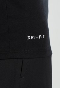 Primer plano de ropa deportiva negra con "DRI-FIT" impreso en blanco cerca del dobladillo, mostrando la textura de la tela y una costura de bolsillo debajo.