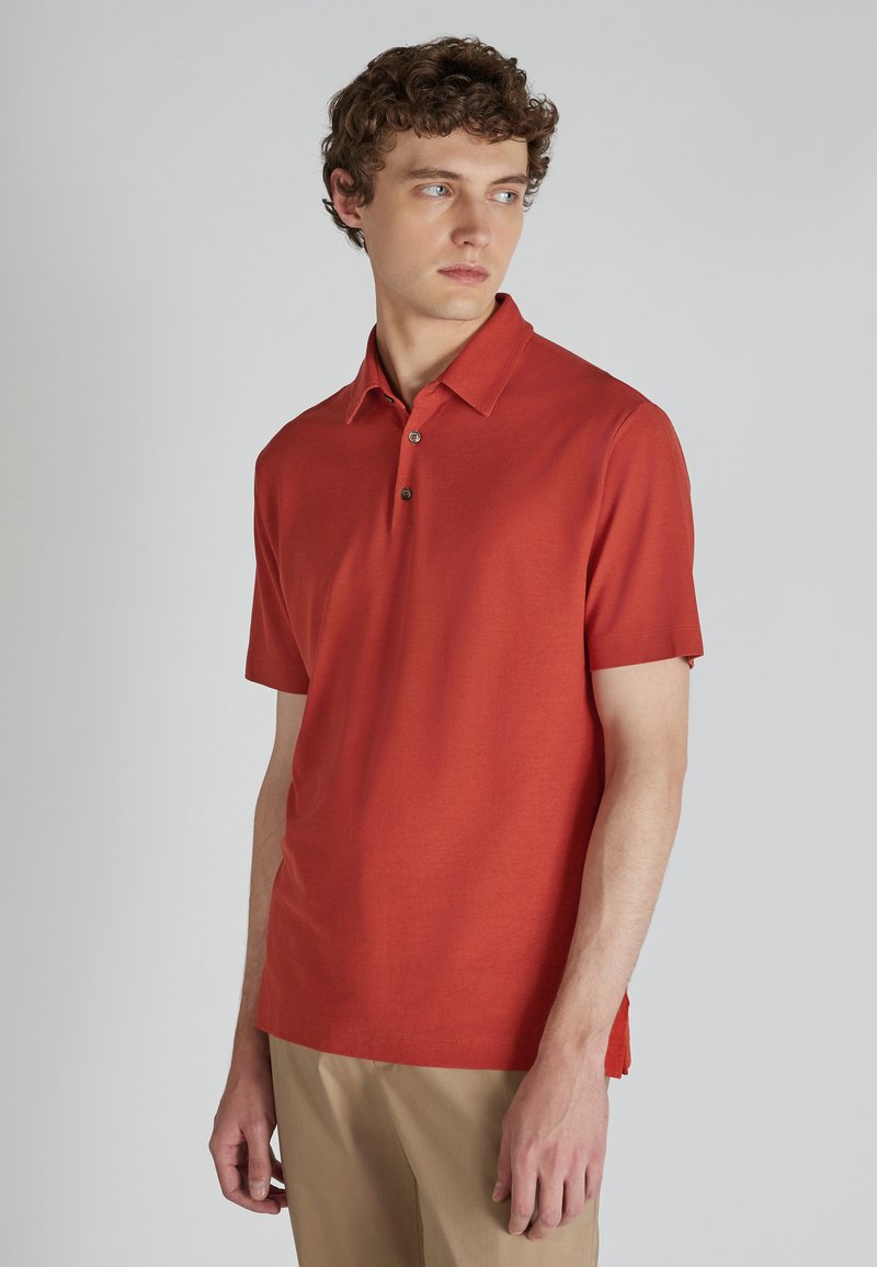 Polo shirt da uomo a maniche corte in tessuto rosso mattone. Presenta un colletto classico, una patta con tre bottoni e fessure laterali per facilitare i movimenti.