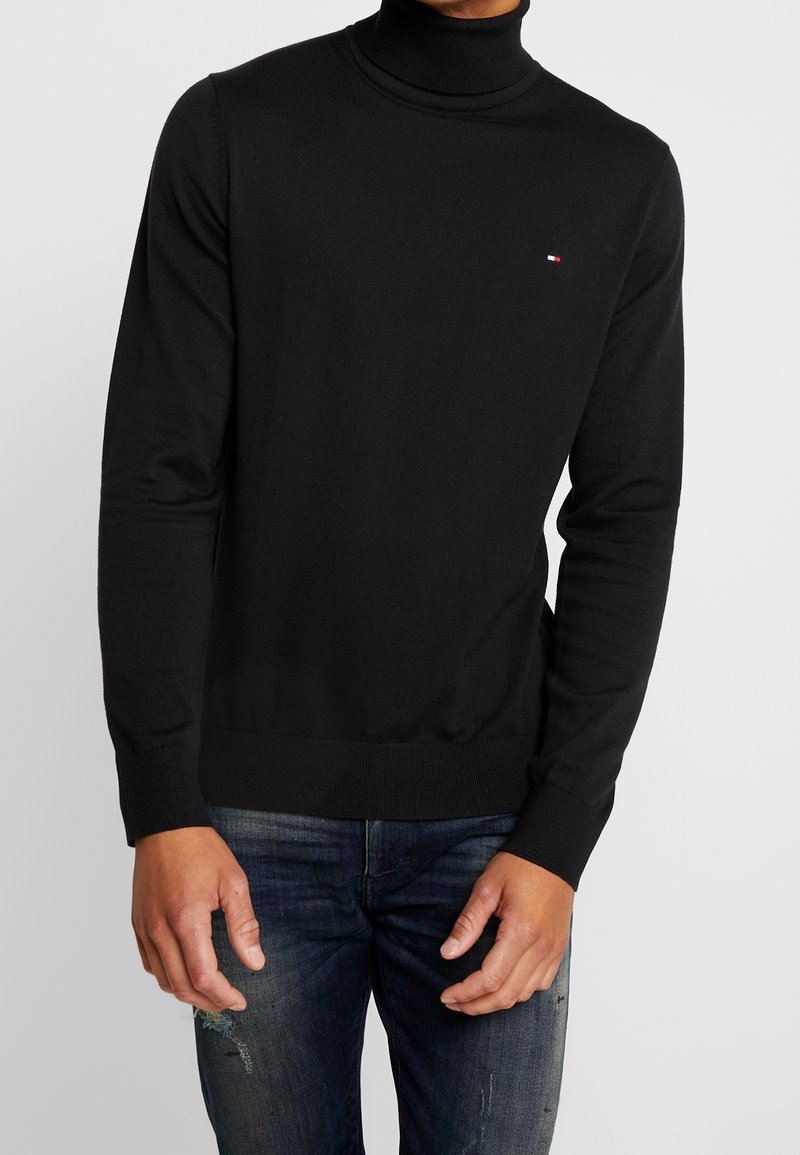 Homme portant un pull à col roulé noir avec un petit logo sur la poitrine et un jean bleu foncé délavé sur fond uni.