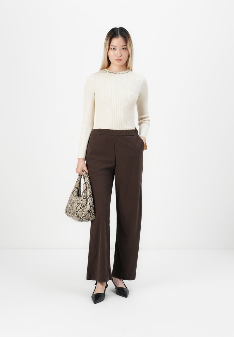 Vrouw in een roomkleurige ribgebreide lange mouw top, bruine wijde broek, zwarte puntige schoenen, met een handtas met slangenprint.