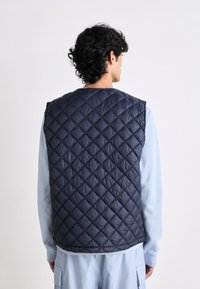 Navy quilted vest with a high collar and sleeveless design, featuring a diamond pattern and a smooth, shiny texture.