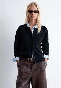 Schwarze Strickjacke mit Knöpfen, gerippten Bündchen, getragen über einem lichtblauen Hemd mit Kragen. Kombiniert mit braunen Lederhosen. Accessoires umfassen eine Sonnenbrille.