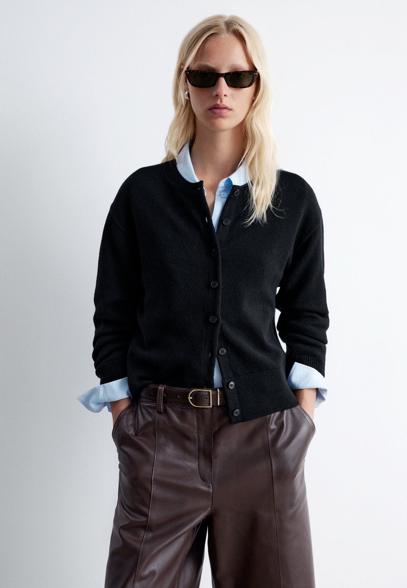 Schwarze Strickjacke mit Knöpfen, gerippten Bündchen, getragen über einem lichtblauen Hemd mit Kragen. Kombiniert mit braunen Lederhosen. Accessoires umfassen eine Sonnenbrille.