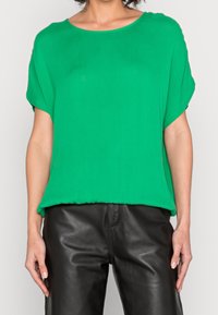 Blouse verte à manches courtes avec une coupe ample, en tissu léger. Elle présente un col rond et tombe légèrement en dessous de la taille.
