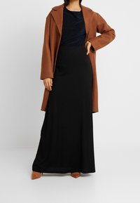 Manteau oversize marron sur un haut bleu marine foncé et une jupe maxi noire, accompagné de chaussures à talons aiguilles beiges. Textures lisses et design superposé.