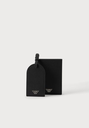 Etiqueta de equipaje y cartera de cuero negro, ambas con el logotipo plateado de águila de Emporio Armani, expuestas una al lado de la otra sobre un fondo blanco liso.