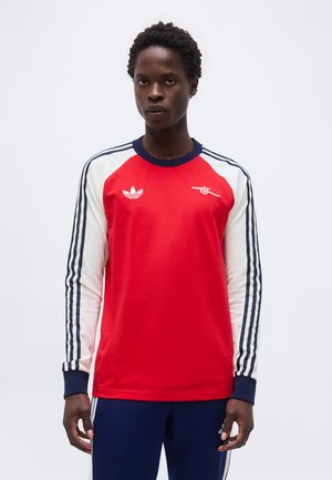 Persona vistiendo una camiseta deportiva de manga larga roja y blanca con los logotipos de Adidas y Arsenal, combinada con pantalones azul marino con rayas blancas.