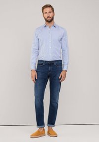 Camicia a maniche lunghe a righe blu e bianche, jeans blu scuro e scarpe gialle senza lacci, in piedi contro uno sfondo neutro.