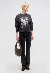 Άτομο με κοντά ξανθά μαλλιά, που φοράει μαύρο φούτερ με βοτανικό σχέδιο και μαύρο τζιν, κρατώντας μια τσάντα χειρός σε υφή λαδί πράσινου χρώματος.