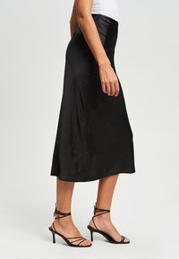 Musta midi-hame, joka on valmistettu sileästä, kiiltävästä kankaasta. Hameessa on istuva vyötärö ja levenevä helma. Yhdistettynä ohuisiin mustiin korkokenkiin.