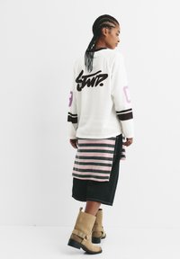 Weißes Langarmhemd mit schwarzem Logo, gestreifter Überwurf in Pink und Grau, kombiniert mit schwarzen Denim-Shorts und beige ankle boots.
