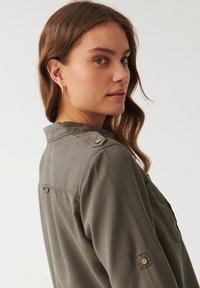 Camisa verde oliva con cuello abotonado, detalles en los hombros y una tela texturada. Mangas largas con costuras decorativas y cierre de botones.