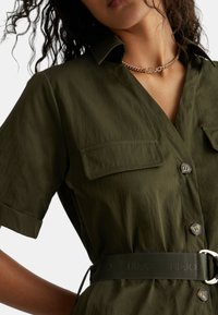 Abito camicia verde oliva con maniche corte, due tasche sul petto, chiusura a bottoni e una cintura con logo dotata di fibbia metallica. Tessuto liscio.