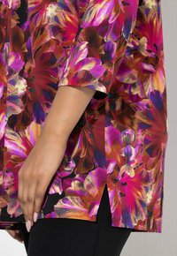 Kwiecista tunika z żywymi różowymi, fioletowymi i pomarańczowymi wzorami na czarnym tle. Charakteryzuje się krótkimi rękawami i bocznym rozcięciem.