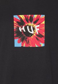 Sort T-shirt med et firkantet motiv, der viser en rød blomst og en blå baggrund, med den hvide tekst "HUF" tydeligt i midten.
