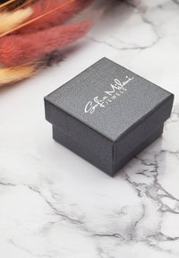 Svart fyrkantig smyckeslåda med en texturerad yta och vitt logo. Ligger mot en marmor bakgrund med mjuka, suddiga pastellfärger.