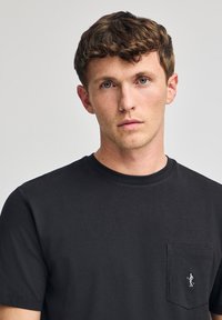 T-shirt in cotone nero con collo girocollo, maniche corte e taschino sul lato sinistro con un piccolo logo ricamato. Texture liscia.