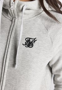 Sudadera con capucha gris con cremallera en tejido suave que presenta un bolsillo canguro y un logo bordado en negro en el pecho izquierdo. Costuras texturizadas a lo largo de las uniones.