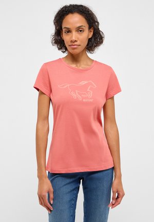 Woman with curly hair wearing a coral pink t-shirt with a white running horse graphic and blue jeans, standing against a plain light background.