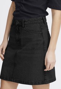 Jupe en denim noir avec une coupe droite, taille mi-haute, poches avant et détails de couture subtils. Le matériau a une finition légèrement texturée.