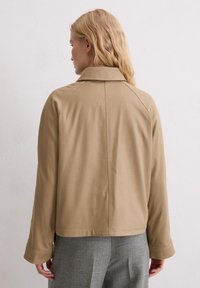 Beżowa kurtka z kołnierzem, wykonana z lekkiego materiału, z szerokimi mankietami. Charakteryzuje się prostym wzorem i widocznym przeszyciem na plecach.