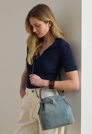 Kobieta o długich blond włosach, ubrana w granatowy prążkowany top i beżowe spodnie, trzymająca jasnoniebieską torebkę z złotym zapięciem, stojąca przy ścianie.